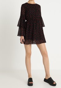 Mujer que lleva un vestido mini negro con estampado de corazones rojos y mangas acampanadas transparentes, combinado con sandalias de plataforma negras.