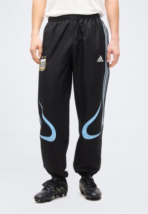 Person trägt schwarze Adidas-Sporthose mit blauen Akzenten, Wappen der argentinischen Fußballmannschaft auf dem linken Oberschenkel und schwarze Stollenschuhe.
