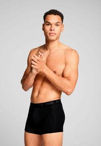 Black boxer shorts with a plain design and elastic waistband featuring a logo. Model is shirtless, displaying a neutral pose.