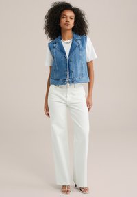 Jeansväst med krage, två framfickor och knappstängning. Bärs över en vit t-shirt och kombineras med vita vida byxor.