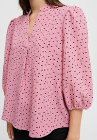 Rosa Bluse mit plissiertem V-Ausschnitt, Puffärmeln und schwarzen Schleifenmustern. Aus leichtem, strukturiertem Stoff gefertigt.