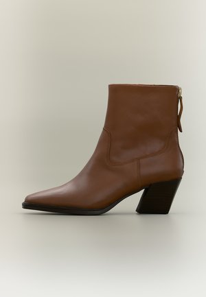 Bottine en cuir marron à bout pointu, talon bloc incliné et fermeture éclair arrière sur fond neutre.