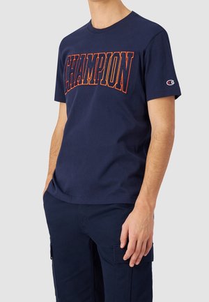 Mand iført marineblå T-shirt med orange "CHAMPION"-tekst og marineblå cargobukser, stående med hænderne i lommerne mod en grå baggrund.