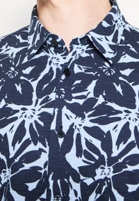 Gros plan d'une chemise bleu marine boutonnée avec un grand motif floral bleu clair sur un tissu texturé.
