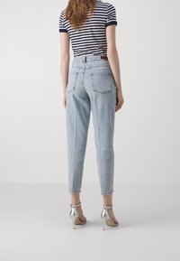 Kvinna med långt hår som bär en randig kortärmad topp, ljusblå högmidjade jeanstights och silversandaler med hög klack, vänd bort.