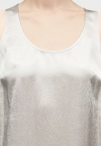 Κοντινή όψη ενός ατόμου που φοράει αμάνικο ασημένιο σατέν τοπ με στρογγυλή λαιμόκοψη, που εμφανίζει τον λαιμό και το πάνω μέρος του στήθους.