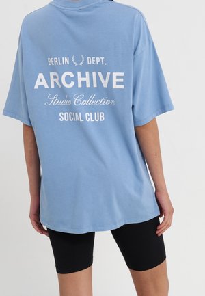 Übergroßes blaues Baumwoll-T-Shirt mit weißem Text: "BERLIN DEPT. ARCHIVE Studio Collection SOCIAL CLUB" auf der Rückseite.