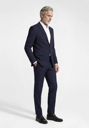 Mann mit grauen Haaren und Bart, der einen dunkelblauen Anzug, weißes Hemd und schwarze Stiefel trägt, steht mit einer Hand in der Tasche vor weißem Hintergrund.