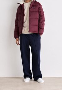 Giacca a vento bordeaux con colletto foderato in pile beige, zip frontale e tasca sul petto. Abbinata a pantaloni blu navy e scarpe bianche.