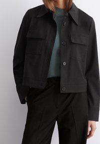 Korte zwarte jas met een knoopsluiting aan de voorkant, twee borstzakken en een klassieke kraag, gecombineerd met een gestructureerde groene top en zwarte broek.