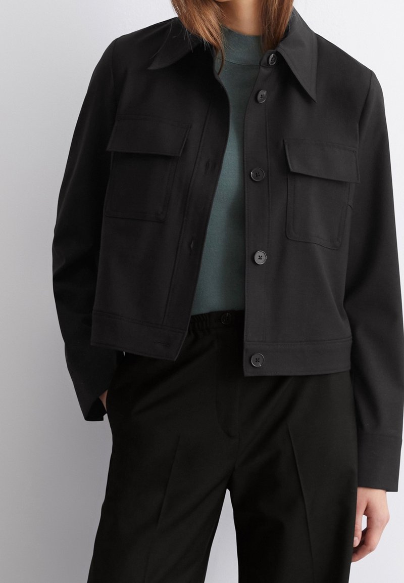 Korte zwarte jas met een knoopsluiting aan de voorkant, twee borstzakken en een klassieke kraag, gecombineerd met een gestructureerde groene top en zwarte broek.