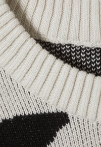 Kötött pulóver bordás krém színű gallérral, fekete és fehér mintákkal, valamint texturált fonallal. Közeli kép a szövet részleteiről és a színek kontrasztjáról.