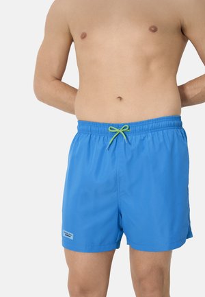 Persona sin camisa que lleva pantalones cortos de baño azul brillante con cordón verde, de pie con las manos detrás de la espalda sobre un fondo blanco.