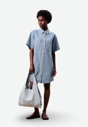 Vrouw draagt een korte mouw spijkerjurk met knopen, houdt een grote lichtgrijze draagtas vast en draagt donkere sandalen met open tenen.