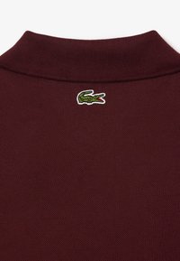 Bordowa koszulka polo z teksturowanej tkaniny, z zielono-białym haftowanym logo krokodyla na kołnierzyku.