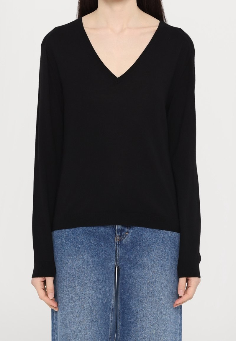 Femme portant un pull noir uni à manches longues en col en V et un jean bleu, debout devant un fond blanc.