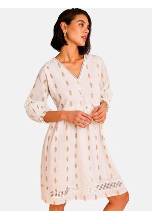 Femme portant une robe blanche longueur genou avec des motifs de feuilles dorées, manches trois-quarts et bordure en dentelle, regardant sur le côté.