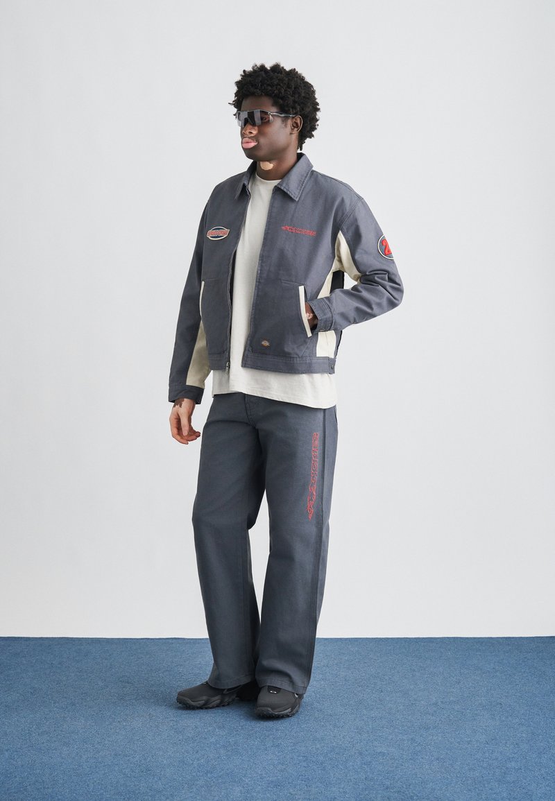 Gray zip-up jacket with cream sleeves, red embroidery, matching gray pants, and black footwear. Model wearing sunglasses. Textured fabric.