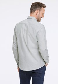 Camicia grigia chiaro con maniche lunghe, colletto classico e dettagli a righe sul retro, abbinata a jeans scuri.