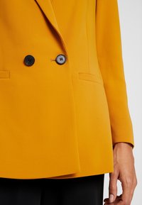 Dwurzędowa marynarka w kolorze musztardowej żółci, wykonana z gładkiej tkaniny, wyposażona w dwa czarne guziki i dwie przednie kieszenie. Zbliżenie klapy i rękawa.