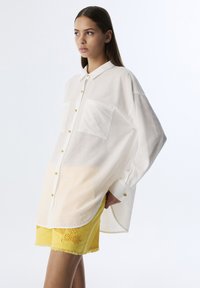 Weißes Hemd mit Knöpfen und Taschen aus strukturiertem Stoff, kombiniert mit leuchtend gelben Shorts mit gesticktem Detail und ausgefranstem Saum.