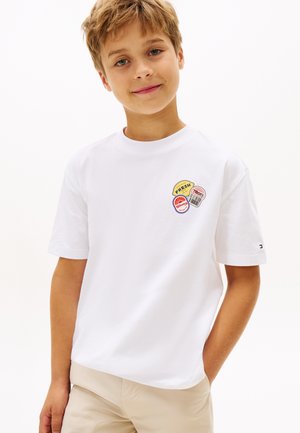 Smilende dreng med lyst brunt hår iført hvid T-shirt med farverige runde pletter og beige bukser, stående foran en ensartet hvid baggrund.