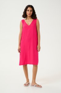Robe sans manches de couleur rose vif avec un décolleté en V, en tissu léger, tombant juste au-dessus des genoux. Portée avec des tongs blanches.