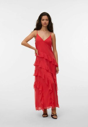 Nő piros ujjatlan maxiruha diagonális fodrokkal, az egyik kezével a csípőjén áll, fekete pántos szandált visel.