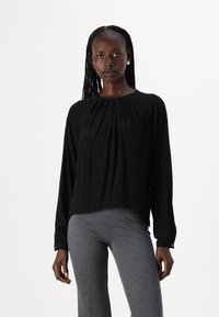 Blusa nera a maniche lunghe con design frontale plissettato, tessuto leggero, scollatura arrotondata e polsini con bottoni, abbinata a pantaloni grigi aderenti.