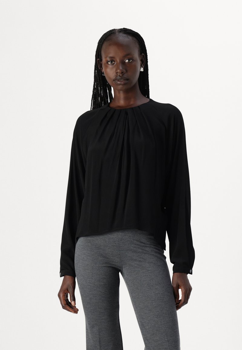 Blusa nera a maniche lunghe con design frontale plissettato, tessuto leggero, scollatura arrotondata e polsini con bottoni, abbinata a pantaloni grigi aderenti.