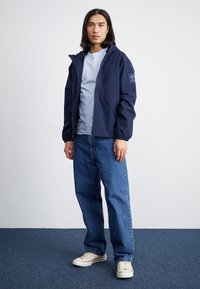 Veste légère marine avec capuche, fermeture éclair à l'avant, deux poches latérales et poignets élastiques, portée sur une chemise bleu clair et un jean bleu.