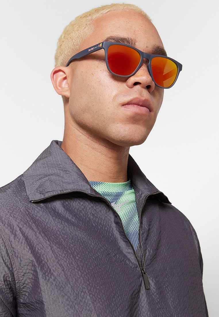 Okulary przeciwsłoneczne z matową, ciemną oprawką i pomarańczowymi lustrzanymi soczewkami. Noszone z kurtką na zamek i teksturowaną, zielono-niebieską koszulą pod spodem.