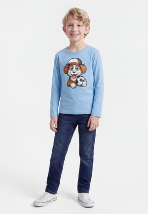 Ragazzo biondo che sorride, indossa una maglia a maniche lunghe celeste con un cane dei cartoni animati che tiene un pallone da calcio, jeans scuri e scarpe da ginnastica bianche.