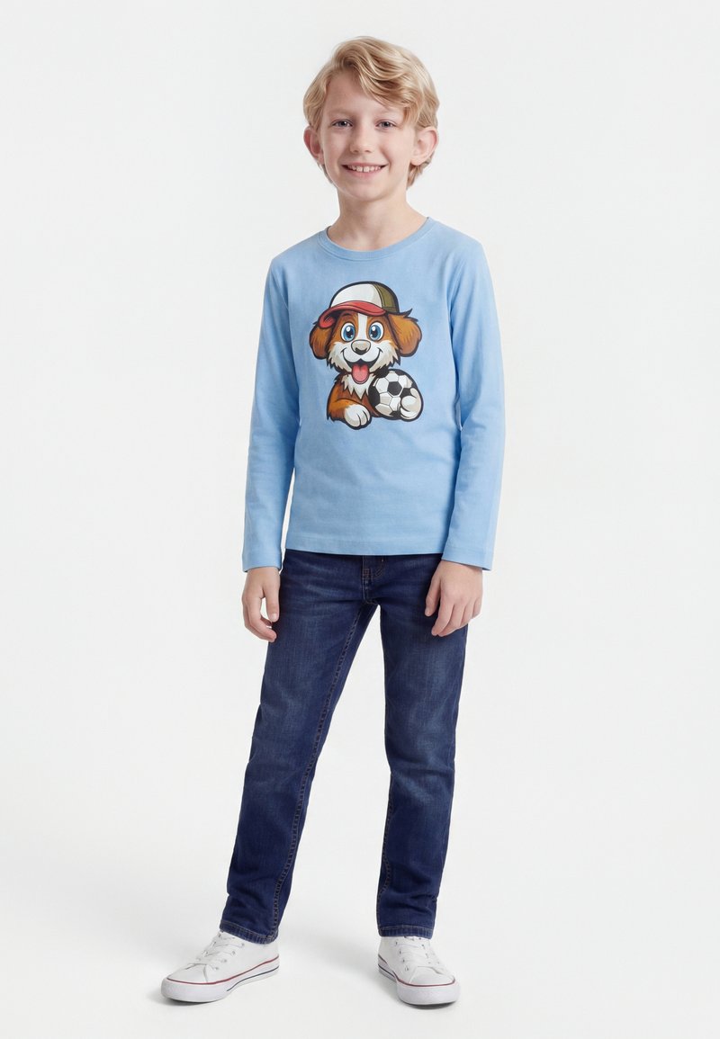 Szőke fiú mosolyog, világoskék hosszú ujjú inget visel, amelyen egy rajzfilm kutya látható, aki focilabdát tart, sötét farmernadrágot és fehér sportcipőt visel.