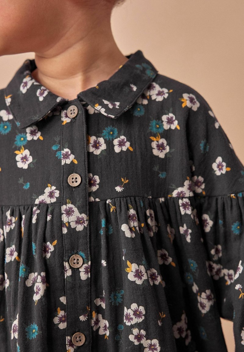 Enfant portant une robe fleurie foncée avec un col et des boutons en bois, visible du cou jusqu'au milieu du torse, regardant sur le côté.