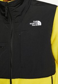 Černo-žlutá vesta s texturovaným, polstrovaným povrchem; přední zip; dvě přední kapsy; s logem The North Face v bílé barvě.