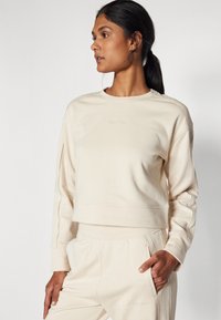 Felpa color crema con scollo rotondo e maniche lunghe, caratterizzata da un ricamo del logo discreto e una vestibilità ampia. Tessuto morbido con una consistenza liscia.