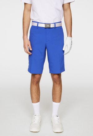 Shorts de golf bleus avec une texture lisse, une taille plate et une coupe droite. Associés à une ceinture blanche, une chemise blanche et des baskets blanches.