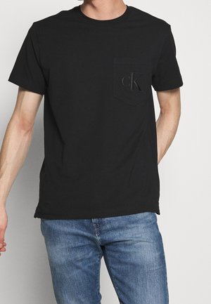 Sort bomuld t-shirt med rund halsudskæring, korte ærmer og en frontlomme med et præget "cK" logo. Båret med blå jeans.