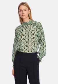 Durchsichtige grüne Bluse mit geometrischem Muster, hoch aufgerichtetem gerafftem Kragen und puffigen Ärmeln. Kombiniert mit maßgeschneiderten dunklen Hosen.