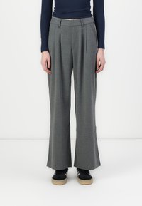 Calças largas cinzentas com pregas frontais, feitas de um tecido texturizado. Combinadas com sapatilhas pretas com sola bege e três riscas brancas.