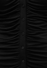 Μαύρο ύφασμα με κάθετη σειρά από πέντε μαύρα κουμπιά, με λεπτομέρεια ραφής με σούρες και στις δύο πλευρές της λωρίδας των κουμπιών.