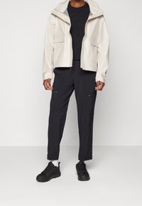 Veste beige clair imperméable avec capuche, fermeture éclair à l'avant, deux grandes poches, associée à un pantalon cargo noir et des baskets noires.