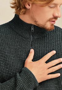 Pulover tricotat de culoare verde închis cu guler înalt și închidere cu fermoar. Prezintă un material texturat și manșete strâmte pentru o ajustare confortabilă.