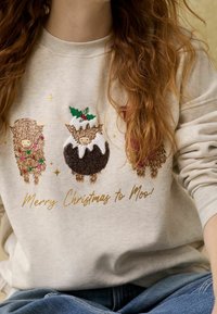 Sweat-shirt gris clair avec trois vaches brodées décorées de motifs festifs, et texte en or disant « Joyeux Noël à Moo ! »
