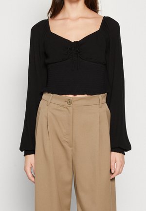 Femme portant un haut court noir froncé à manches longues avec un décolleté carré et un pantalon beige plissé taille haute.
