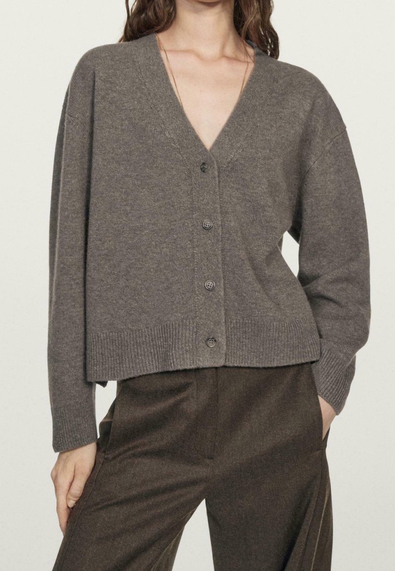 Femme portant un cardigan gris ample boutonné avec un col en V et un pantalon marron foncé, une main dans la poche et l'autre reposant à côté.