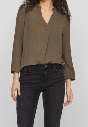 Blouse verte olive à manches longues avec un col en V, en tissu léger et avec des poignets légèrement roulés, associée à un jean noir ajusté.