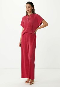 Červený plisovaný top s krátkým rukávem a výstřihem do V, spárovaný s širokými červenými kalhotami. Hladká tkanina s jemnou texturou. Zlaté sandály.