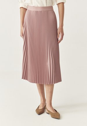 Femme portant une jupe midi plissée rose poudré, un haut blanc et des chaussures en daim marron à talons bas et bouts pointus, debout devant un fond uni.
